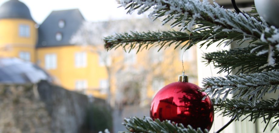 Nahaufnahme eines geschmückten Tannenzweigs mit roter Kugel und Schneeeffekt, im Hintergrund das gelbe Schloss Montabaur mit Türmen und grauen Dächern, leicht verschwommen