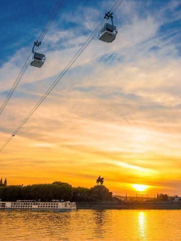 Panoramaaufnahme eines Flusses bei Sonnenuntergang, zwei Seilbahngondeln am Himmel, ruhige Wasseroberfläche mit Spiegelungen, Ufer mit Bäumen und Denkmal im Hintergrund, Himmel in warmen Gelb- und Orangetönen