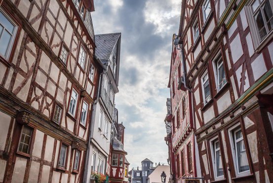 Schmale Kopfsteinpflasterstraße in Montabaur, gesäumt von hohen Fachwerkhäusern mit roten und weißen Balken, Blumendekoration an Fenstern, Menschen flanieren zwischen Geschäften, im Hintergrund ein heller Himmel mit Wolken