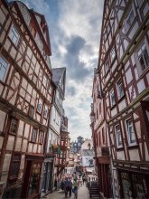 Historische Altstadt von Montabaur – Fachwerkcharme und lebendige Gassen Schmale Kopfsteinpflasterstraße in Montabaur, gesäumt von hohen Fachwerkhäusern mit roten und weißen Balken, Blumendekoration an Fenstern, Menschen flanieren zwischen Geschäften, im Hintergrund ein heller Himmel mit Wolken