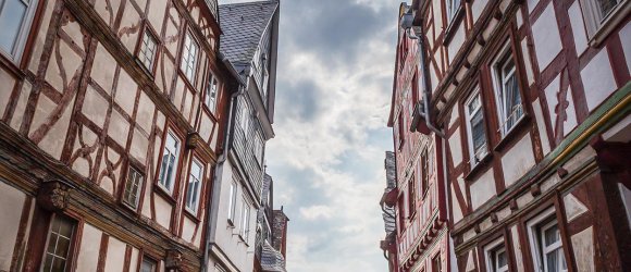Historische Altstadt von Montabaur – Fachwerkcharme und lebendige Gassen Schmale Kopfsteinpflasterstraße in Montabaur, gesäumt von hohen Fachwerkhäusern mit roten und weißen Balken, Blumendekoration an Fenstern, Menschen flanieren zwischen Geschäften, im Hintergrund ein heller Himmel mit Wolken