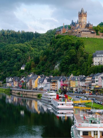 Panoramaaufnahme eines ruhigen Flusses mit mehreren weißen Ausflugsschiffen am Ufer, farbenfrohen Häusern entlang der Promenade und einem großen Schloss auf einem bewaldeten Hügel, bewölkter Himmel im Hintergrund
