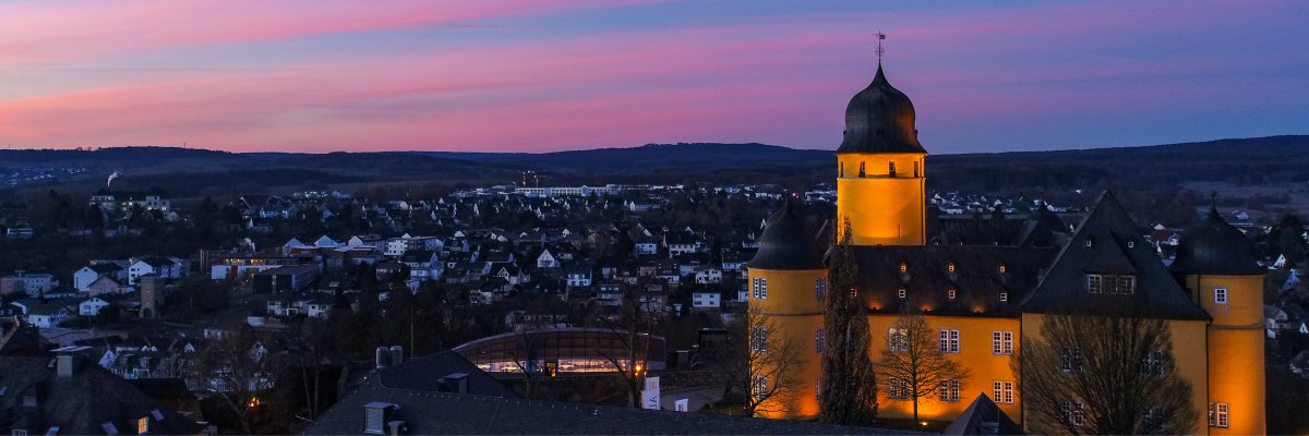 Panoramaaufnahme eines gelb beleuchteten Schlosses mit markantem Turm, umgeben von Häusern und sanften Hügeln, Himmel in Rosa- und Blautönen während der Dämmerung