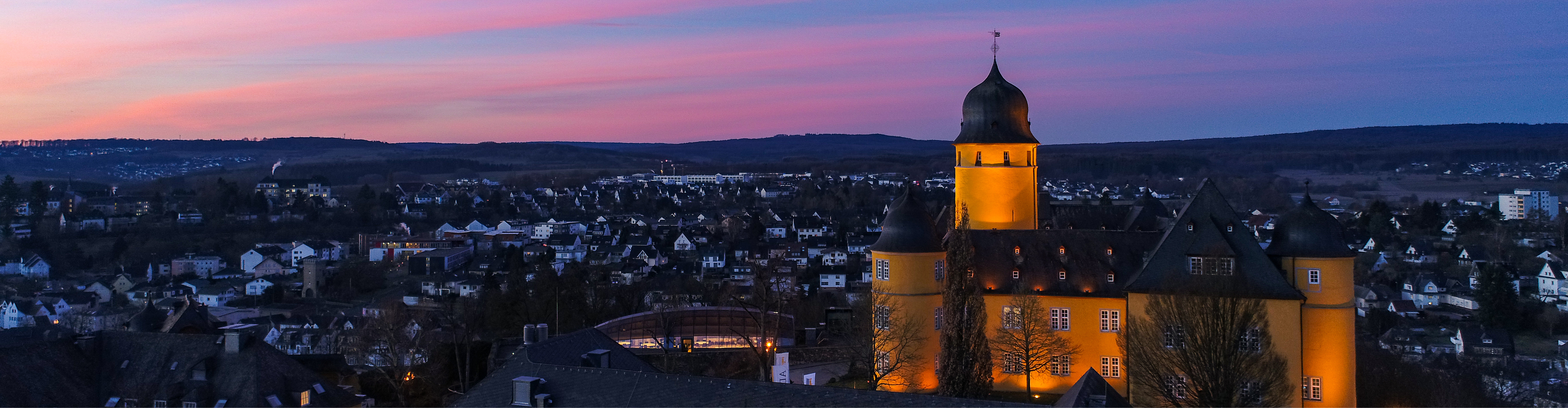 Panoramaaufnahme eines gelb beleuchteten Schlosses mit markantem Turm während der Dämmerung