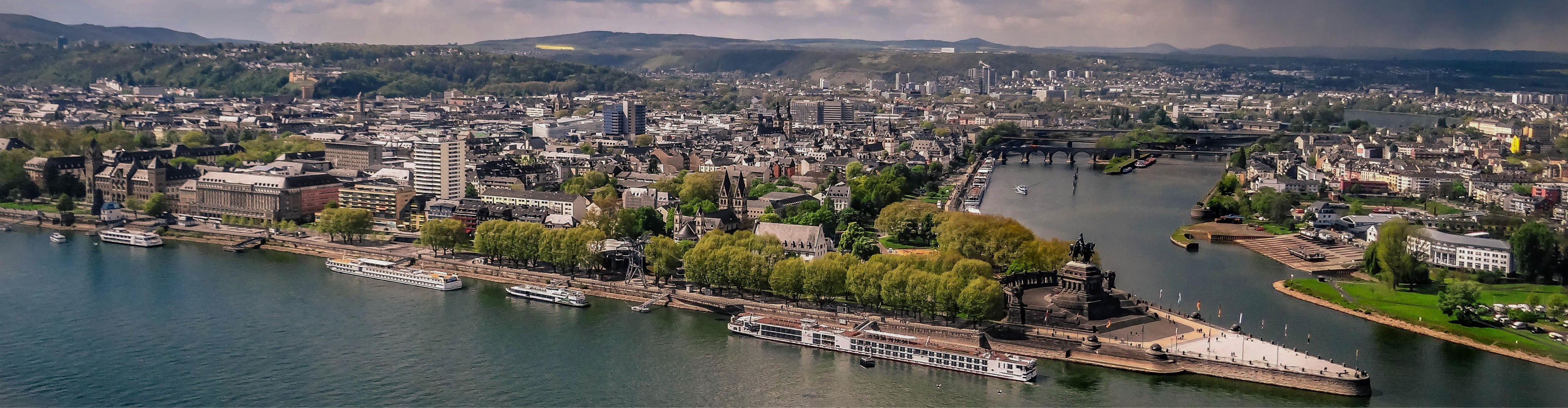 Wide-angle image of Koblenz featuring the Deutsches Eck, several white excursion boats along the shore, a broad promenade lined with trees, bridges crossing the river, and an expansive city in the background surrounded by gentle hills under a cloudy sky
