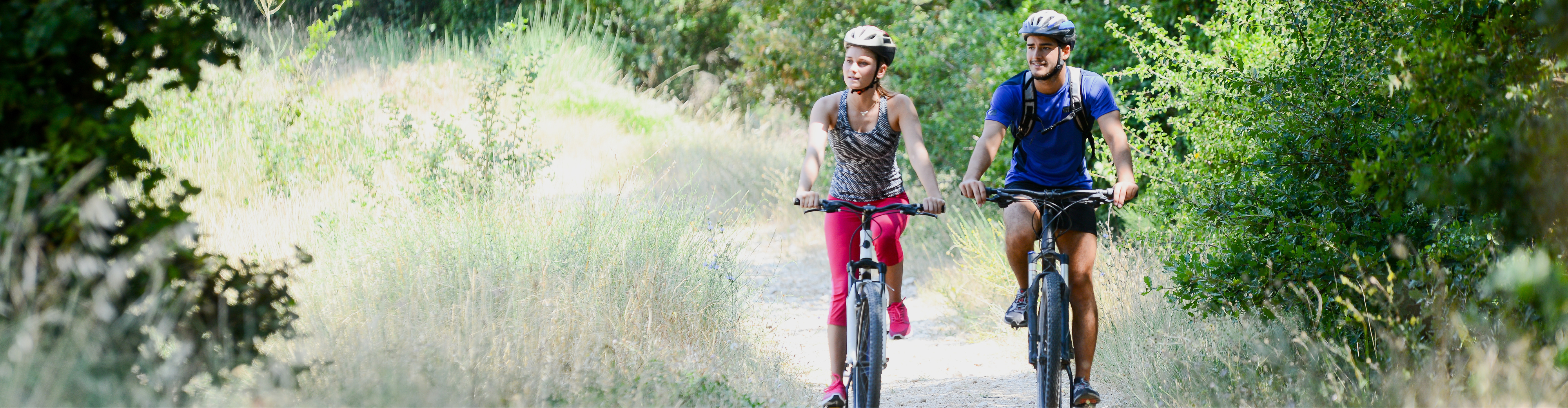 Active in Nature – Cycling Tour on a Scenic Forest Path Two cyclists on a narrow, bright forest trail surrounded by dense greenery and tall shrubs, sunlight filtering through trees creating bright patches on the ground