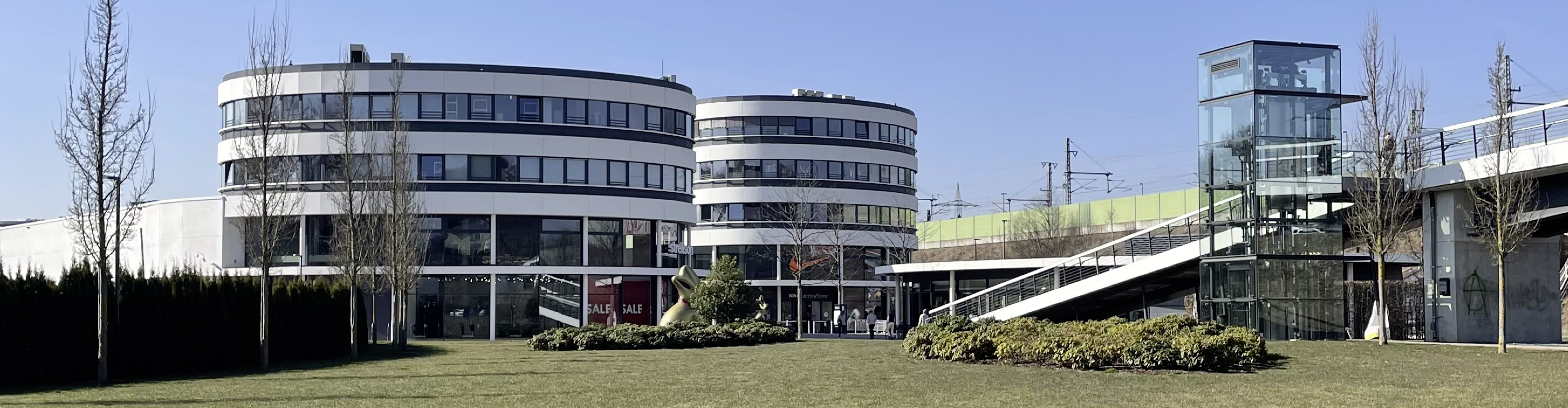 Außenansicht des Fashion Outlet Center Montabaur mit zwei runden Gebäuden in Weiß und Glas, gläsernem Aufzugsturm, breiter Rampe, gepflegtem Rasen und jungen Bäumen, im Hintergrund Bahngleise und Oberleitungen unter klarem Himmel