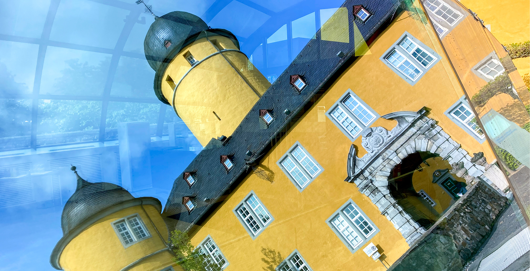 Schloss Montabaur – Historische Architektur in modernem Blickwinkel Gelbes Schloss Montabaur mit Türmen und grauen Dächern, durch Glas fotografiert mit Spiegelungen und blauen Lichtreflexen, im Vordergrund der barocke Torbogen mit Steinverzierungen