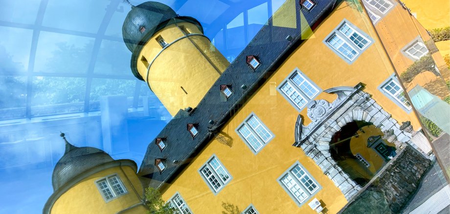 Gelbes Schloss Montabaur mit Türmen und grauen Dächern, durch Glas fotografiert mit Spiegelungen und blauen Lichtreflexen, im Vordergrund der barocke Torbogen mit Steinverzierungen