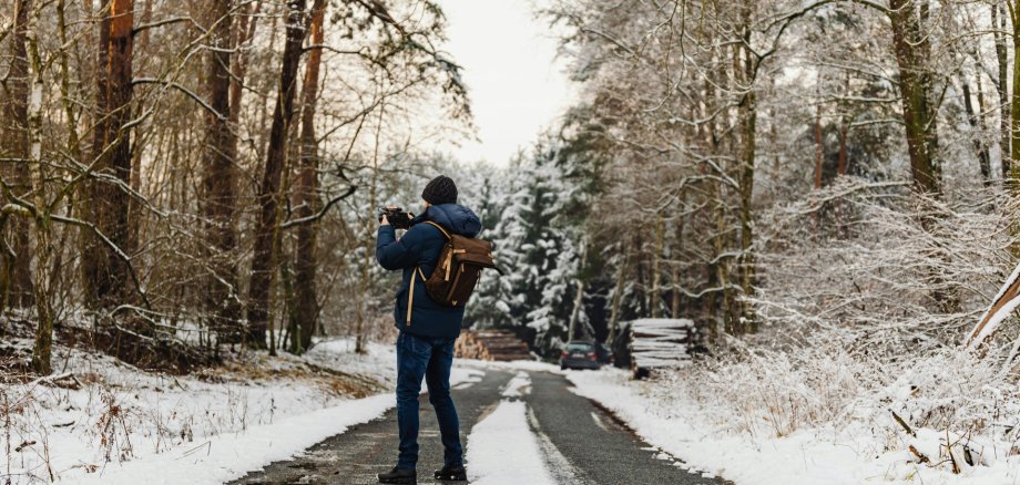 Person steht auf einem schmalen Waldweg, umgeben von verschneiten Bäumen, trägt Winterjacke, Mütze und Rucksack, fotografiert die winterliche Landschaft unter hellem Himmel
