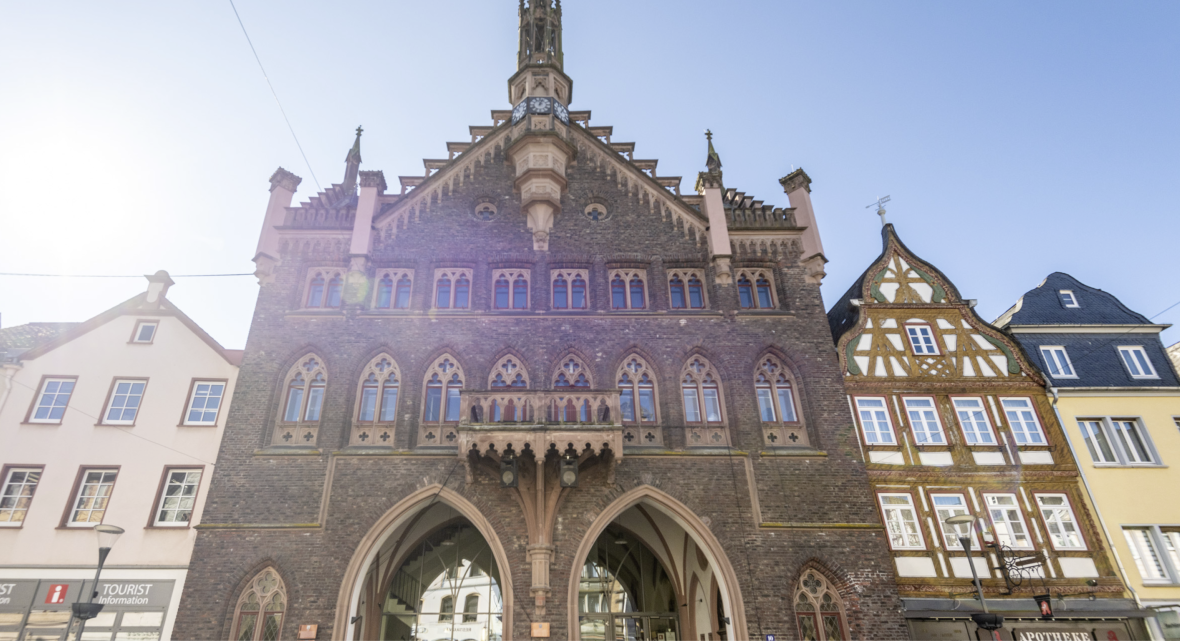 Historisches Rathaus in Montabaur – Architektur mit Geschichte Frontansicht des historischen Rathauses in Montabaur mit dunkler Steinfassade, Spitzbogenarkaden, verzierten Fenstern und Turmspitze, umgeben von hellen Gebäuden und einem Fachwerkhaus, blauer Himmel im Hintergrund