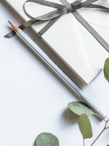 Nahaufnahme eines weißen Geschenkgutscheins mit grauem Satinband und Schleife, daneben ein silberner Bleistift und grüne Eukalyptusblätter auf hellem Untergrund