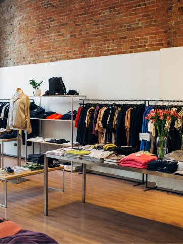 Innenansicht eines modernen Modegeschäfts mit Backsteinwand, Kleiderstangen voller farbiger Kleidung, Regalen mit gefalteten Shirts und Hosen, einem Tisch mit Accessoires sowie einer Vase mit Blumen