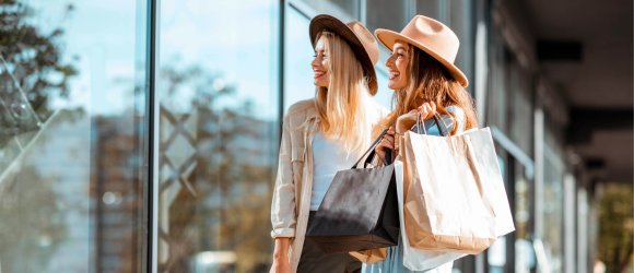 Shopping im Fashion Outlet Center Montabaur Zwei Personen stehen vor einer Glasfassade mit Einkaufstaschen in der Hand, beide tragen helle Kleidung und Hüte, Spiegelungen der Umgebung im Glas