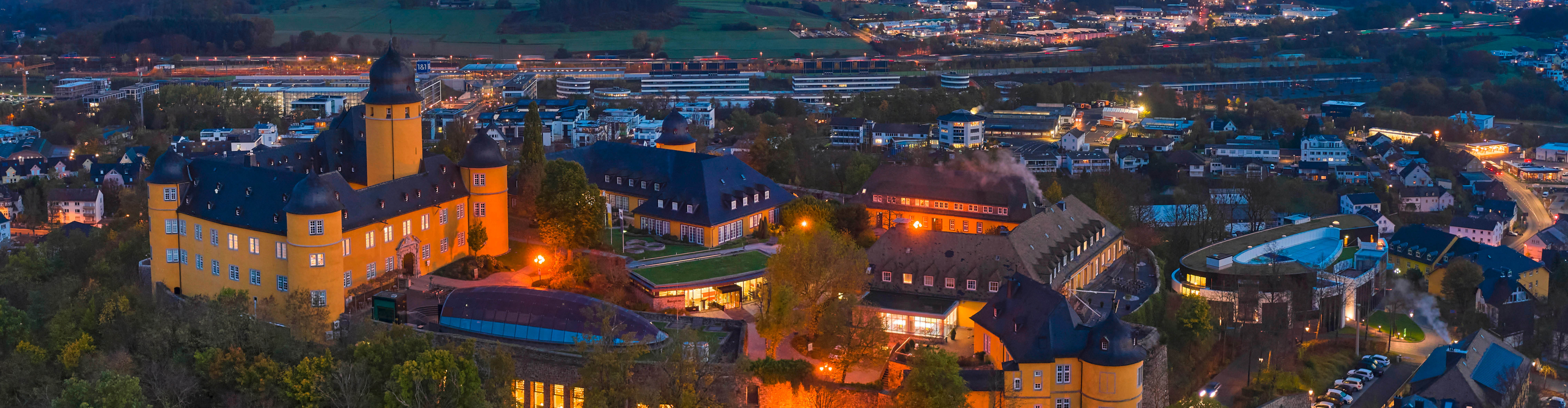 Schloss Montabaur bei Abenddämmerung – Ein Ort voller Geschichte und Moderne Panoramaaufnahme von Schloss Montabaur mit gelber Fassade und dunklen Dächern, umgeben von modernen Gebäuden und Straßen, warmes Licht in den Innenhöfen, im Hintergrund die Stadt mit beleuchteten Häusern und Bahntrassen unter blauem Abendhimmel