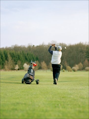 Golf – Relaxation and precision on the green Person swinging a golf club on a large green field, next to a golf trolley with a bag of clubs, with trees and a blue sky in the background