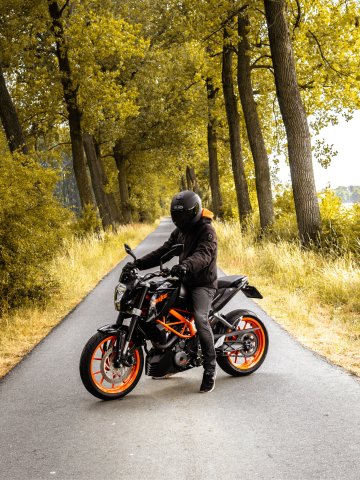 Schwarzes Motorrad mit orangefarbenen Felgen steht auf einer schmalen, asphaltierten Straße, die von hohen Bäumen gesäumt ist, umgeben von grünem Gras und dichter Vegetation
