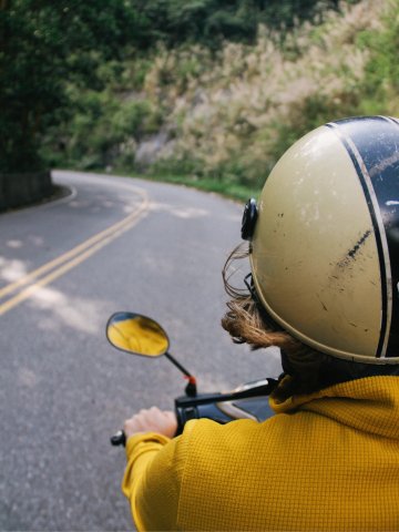 Person mit gelbem Helm und gelber Jacke fährt auf einem Motorrad eine kurvige Landstraße entlang, umgeben von grüner Vegetation und Felsen