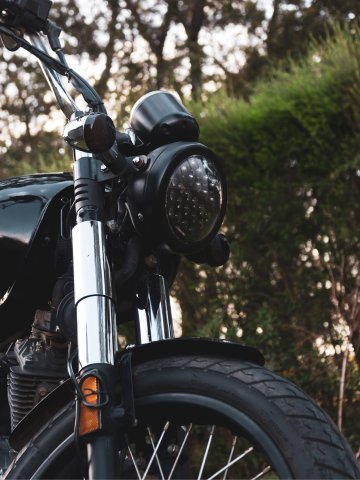 Nahaufnahme eines schwarzen Motorrads mit glänzenden Chromteilen, rundem Frontscheinwerfer und breiten Reifen, im Hintergrund grüne Büsche und Bäume