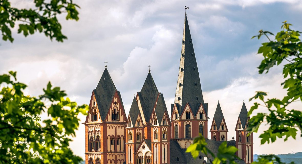 Limburg Cathedral – striking church with red and white facade and tall spires, framed by green foliage under a partly cloudy sky during an excursion from Schloss Montabaur