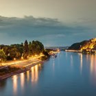 Abendstimmung am Rhein mit Blick auf die Festung Ehrenbreitstein Abendliche Rheinlandschaft – ruhiges Wasser mit Spiegelungen der Lichter, links Uferpromenade mit Bäumen und Gebäuden, rechts die beleuchtete Festung Ehrenbreitstein auf einem Hügel unter wolkigem Himmel