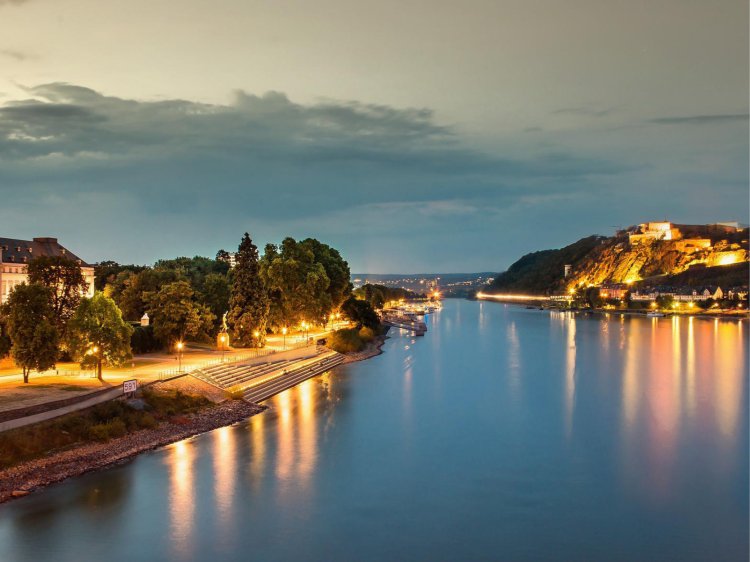 Abendstimmung am Rhein mit Blick auf die Festung Ehrenbreitstein Abendliche Rheinlandschaft – ruhiges Wasser mit Spiegelungen der Lichter, links Uferpromenade mit Bäumen und Gebäuden, rechts die beleuchtete Festung Ehrenbreitstein auf einem Hügel unter wolkigem Himmel