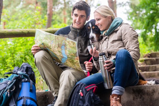 Pause beim Wandern im Westerwald nahe Schloss Montabaur Wandern im Westerwald – zwei Personen sitzen auf einer Steintreppe im Wald, studieren eine Wanderkarte und trinken aus einer Thermoskanne, neben ihnen ein Rucksack und ein Hund, umgeben von grünem Laub