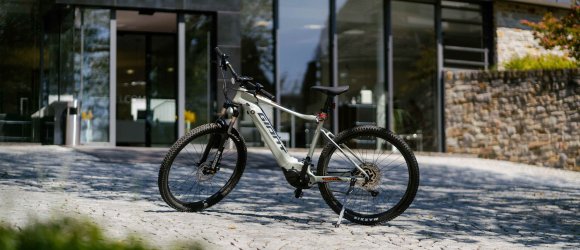 E-Bike vor dem Hotel Schloss Montabaur – Startklar für Ihre Tour Weißes E-Bike der Marke Haibike steht auf einem gepflasterten Vorplatz vor dem modernen Eingangsbereich des Hotels Schloss Montabaur, umgeben von Glasfassade und Natursteinmauer