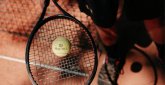 Tennis im Freien Nahaufnahme eines Tennisschlägers mit gelbem Tennisball auf dem Schlägerkopf, darunter roter Sandbelag eines Tennisplatzes, seitlich die Sportschuhe einer Person sichtbar