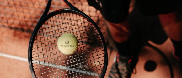 Tennis im Freien Nahaufnahme eines Tennisschlägers mit gelbem Tennisball auf dem Schlägerkopf, darunter roter Sandbelag eines Tennisplatzes, seitlich die Sportschuhe einer Person sichtbar