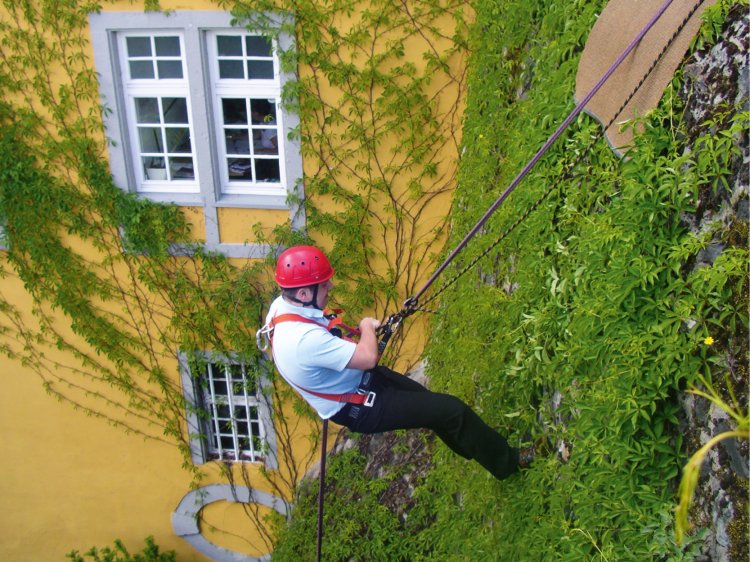 Adventurous Supporting Program – Rappelling at Hotel Schloss Montabaur Individual wearing climbing gear and a red helmet rappelling down a green-covered castle wall in front of a yellow façade with white windows