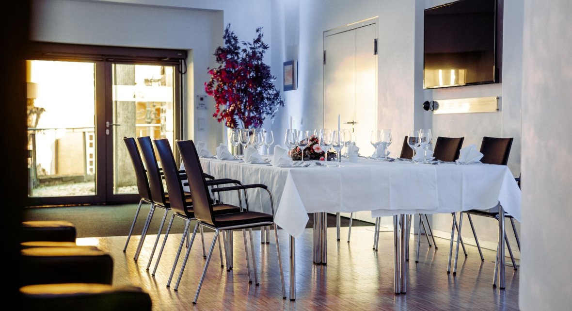 Innenbereich des Veranstaltungszentrums mit weiß gedecktem Tisch, mehreren Stühlen, dekorativen Gläsern und Servietten, großer Leinwand an der Wand und Glastüren mit Blick ins Freie