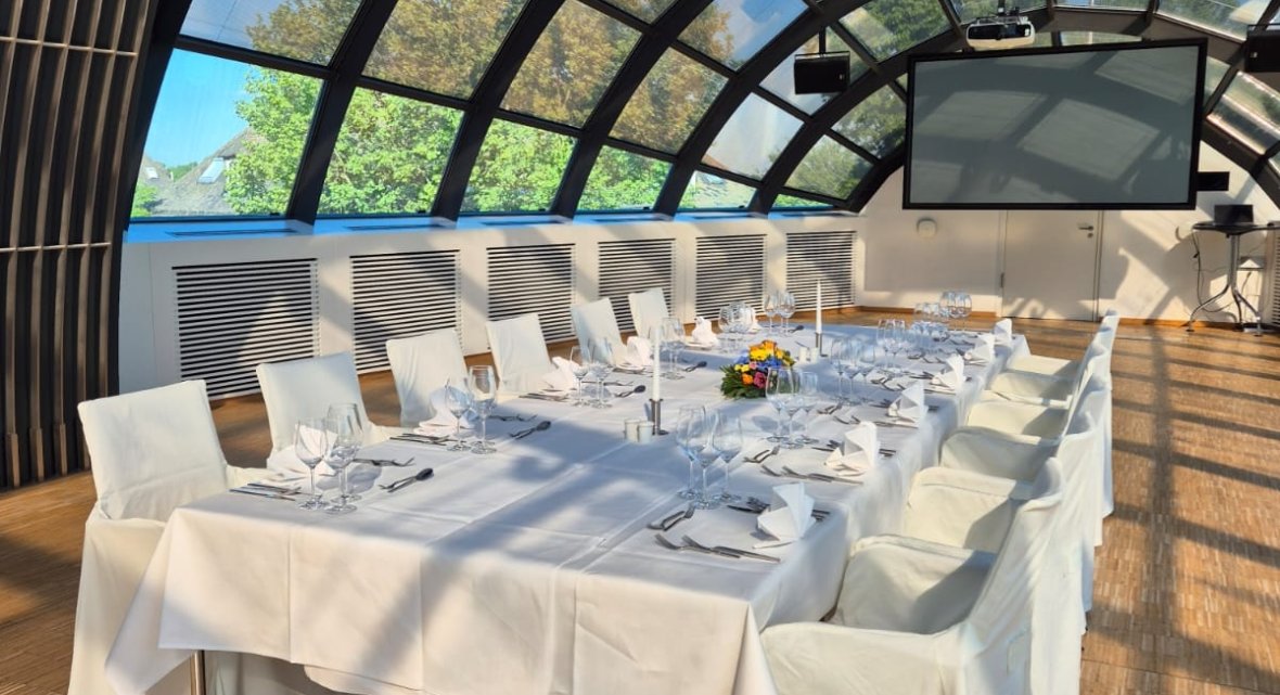 Innenbereich der Kuppel im Veranstaltungszentrum mit großer Glasdachkonstruktion, weiß eingedecktem Tisch, Stühlen mit Hussen, dekorativen Gläsern und einer großen Leinwand an der Stirnseite