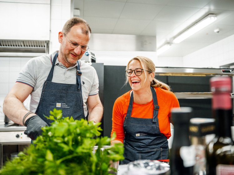 Zwei Personen in Kochschürzen arbeiten in einer modernen Küche, frische Kräuter im Vordergrund, Edelstahlgeräte und Flaschen mit Öl im Hintergrund