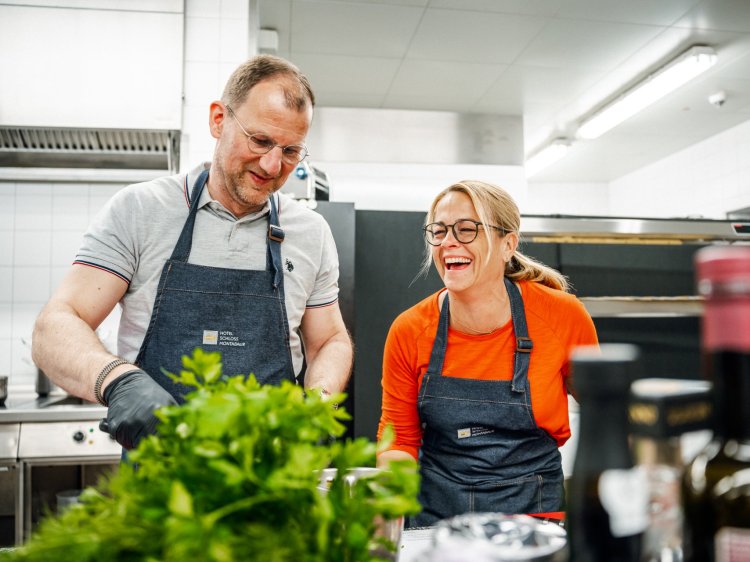 Zwei Personen in Kochschürzen arbeiten in einer modernen Küche, frische Kräuter im Vordergrund, Edelstahlgeräte und Flaschen mit Öl im Hintergrund