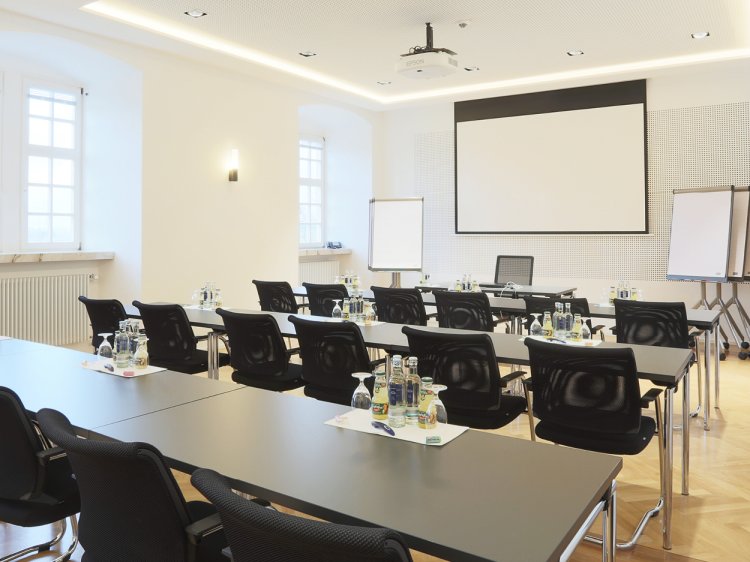 Tagungsraum im Hotel Schloss Montabaur – Moderne Ausstattung für erfolgreiche Meetings Konferenzraum mit mehreren schwarzen Stühlen und Tischen, Wasserflaschen und Gläsern, großer Leinwand an der Stirnseite, Deckenprojektor und Flipcharts, Parkettboden und weiße Wände mit hohen Fenstern