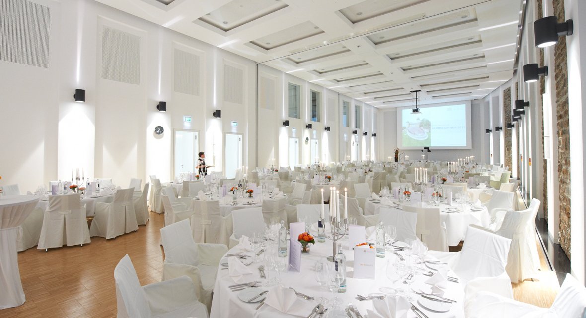 Interior view of a spacious hall featuring multiple round tables with white tablecloths and chair covers, candle holders, orange floral arrangements, cutlery and glasses, bright walls with natural stone accents, wooden flooring, and a projection screen in the background